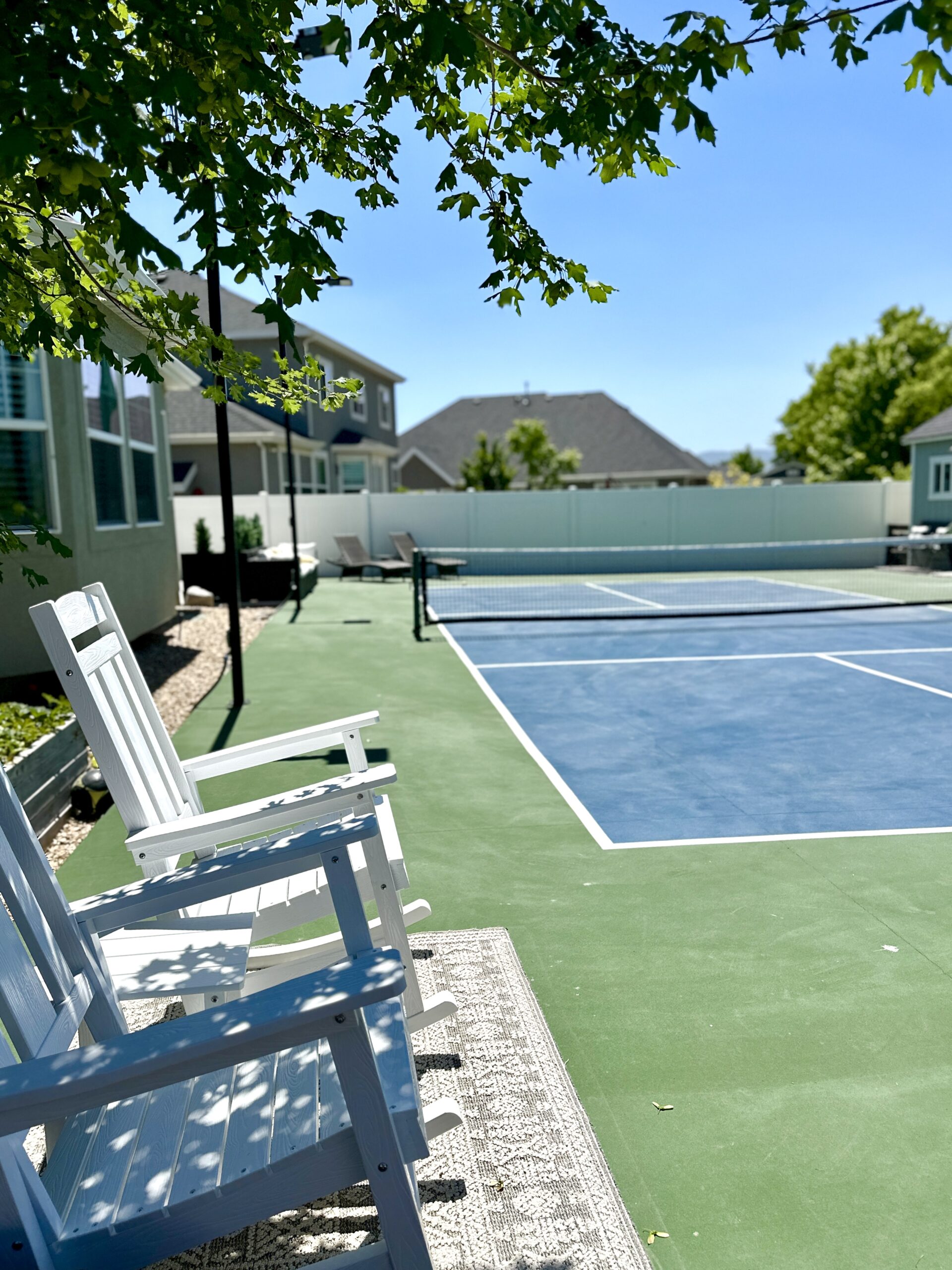 White rocking chairs overlooking a freshly painted backyard pickleball court with blue playing surface.